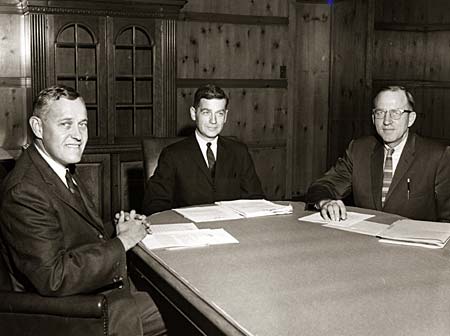 Three men sit at a table with papers in front of them. 