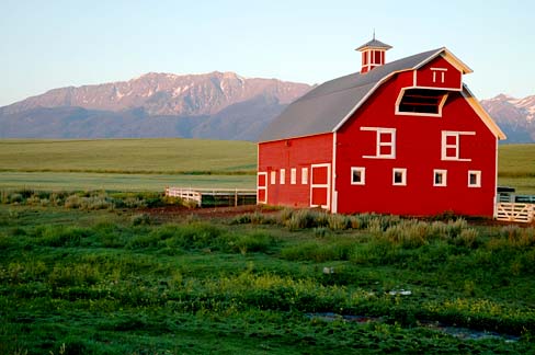 3 story red barn with bell tower. White picket fence on green pasture with snow tipped mountains in back.