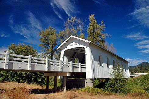 A wooden covered bridge with white guardrail on road leading up to it. Blue sky with a few whispy clouds.