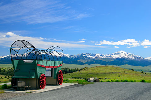 A replica covered wagon stands to the side of a highway with snow capped mountains in the distance.