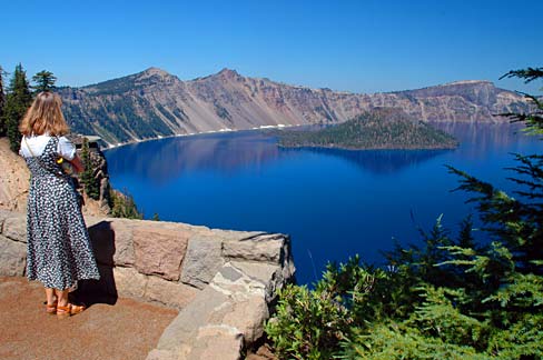Blonde woman in print summer dress stands at overlook area looking out over Crater Lake at Wizard Island.