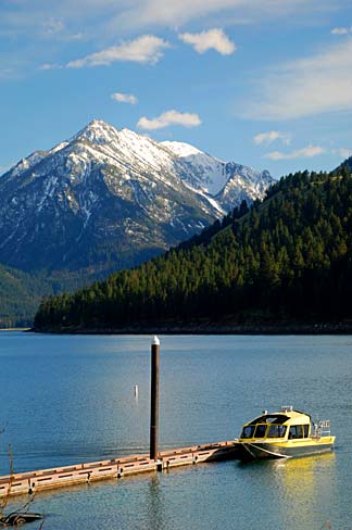 Boat docked at a walkway with snow capped mountain in background. The near shore is covered in evergreen trees.