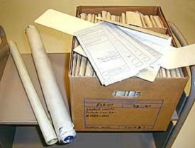 A box on a table with folded files inside and large rolled up, long papers to the side (probably maps).
