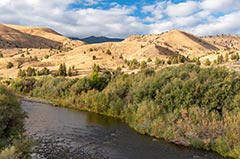 A wide river bordered by dense green shrubs and trees flows through a valley, with rolling golden hills and partly cloudy blue sky in the background..