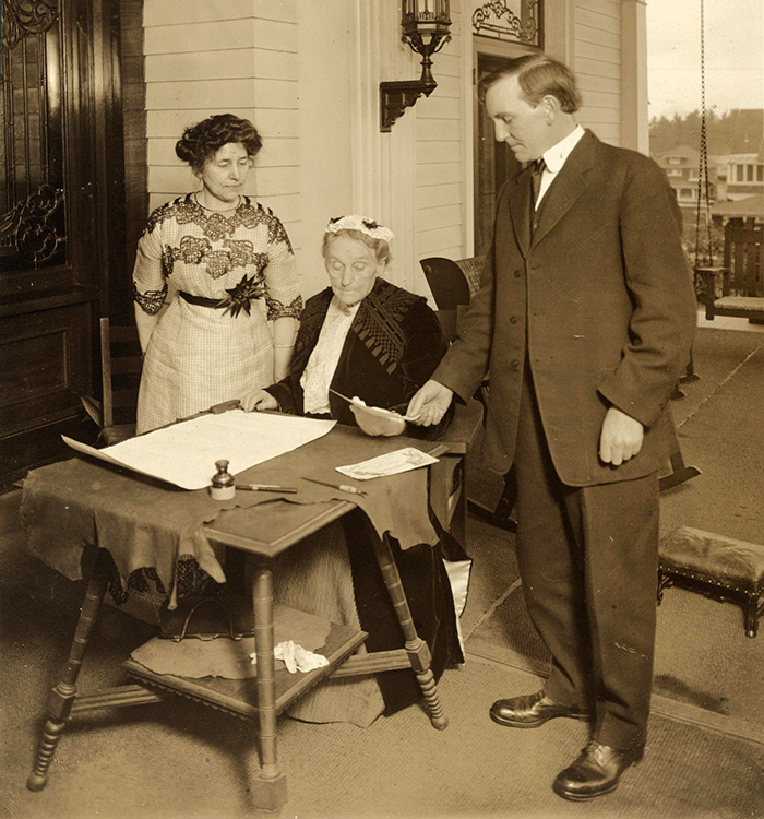 Abigail Scott Duniway sits at a wood table with the proclamation in front of her as she gets ready to sign.