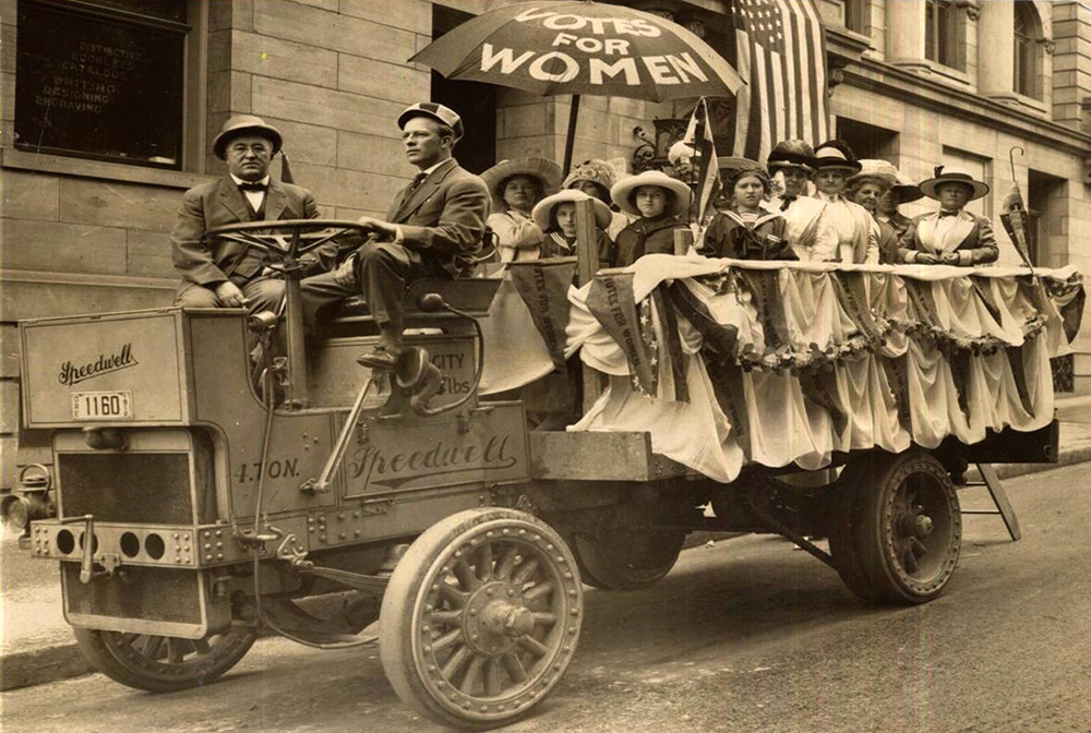 About a dozen women in the back of an open trailer driving by 2 men. Their umbrella reads Votes for Women.