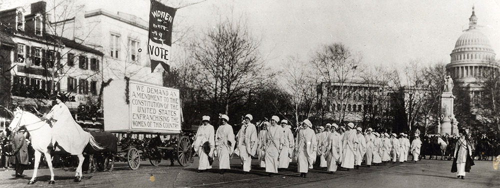 Photograph from 1913. Woman on white horse leads a formation of about 50 women through a street. The capitol building is in the background.