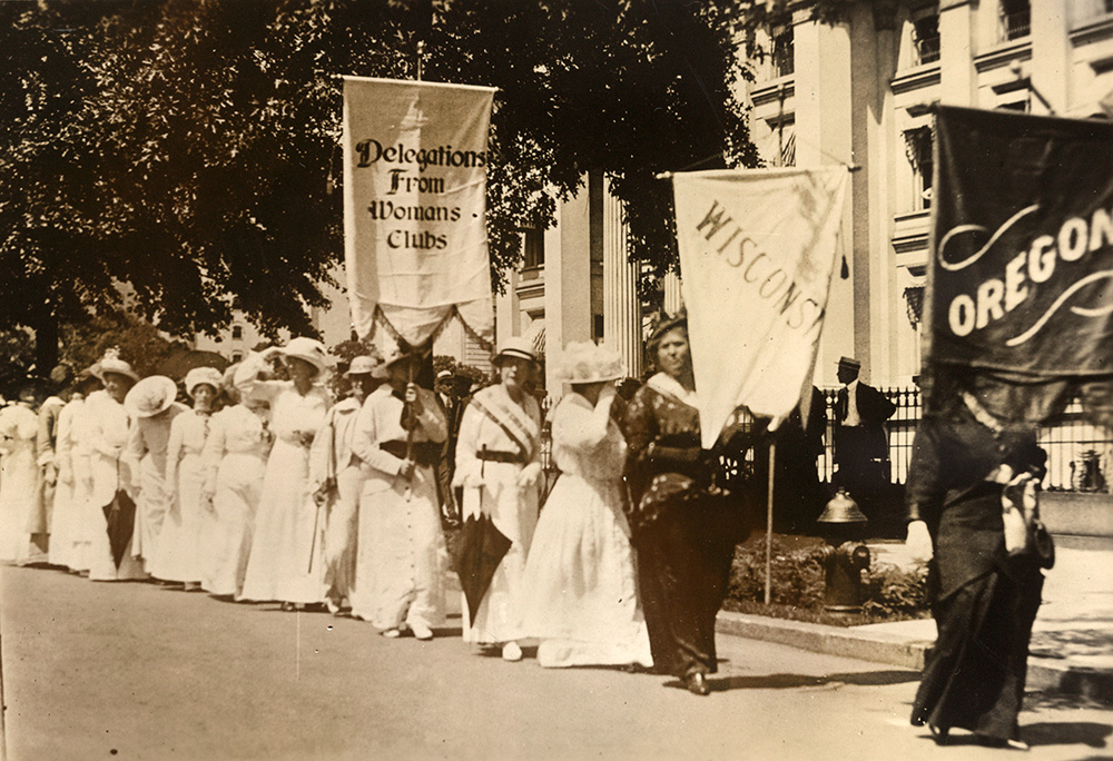 A line of about a dozen women all dressed in white follow behind 2 women in brown and black clothing.