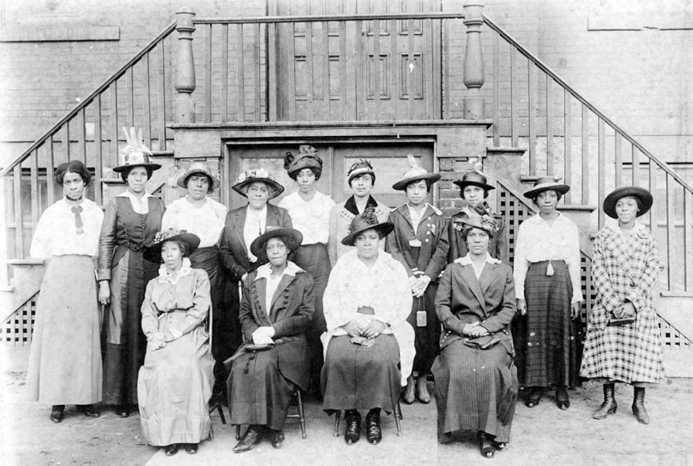 14 women of color pose before a staircase leading to a 2nd story entry door to a building.