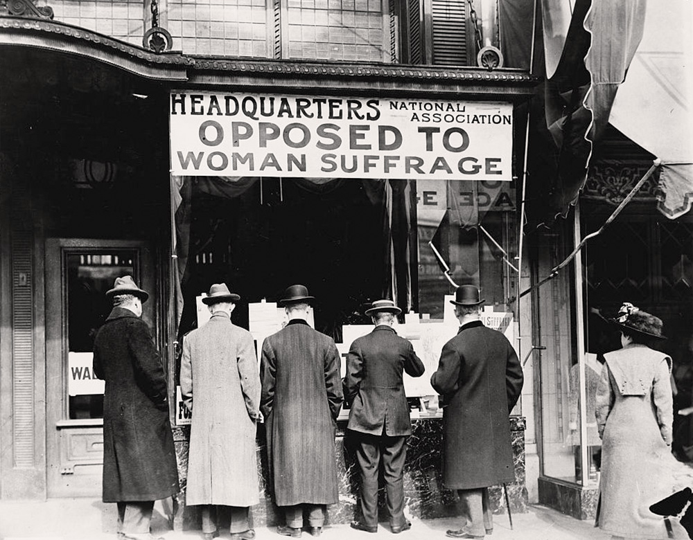 5 men in suits and hats stand with their backs to the camera. They stand in front of the Headquarters of the National Association Opposed to Woman Suffrage.