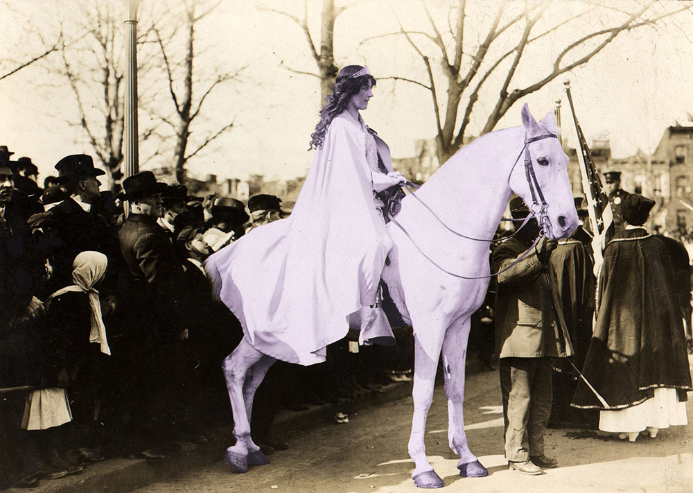 An elegant woman in a long white robe with flowing hair sits on a horse. She is surrounded by a crowd of people standing about.