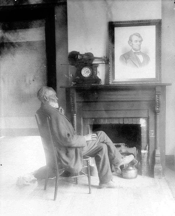 Louis Southworth sits in a straightback wood chair near a fireplace. a picture of Abraham Lincoln hangs on the wall.