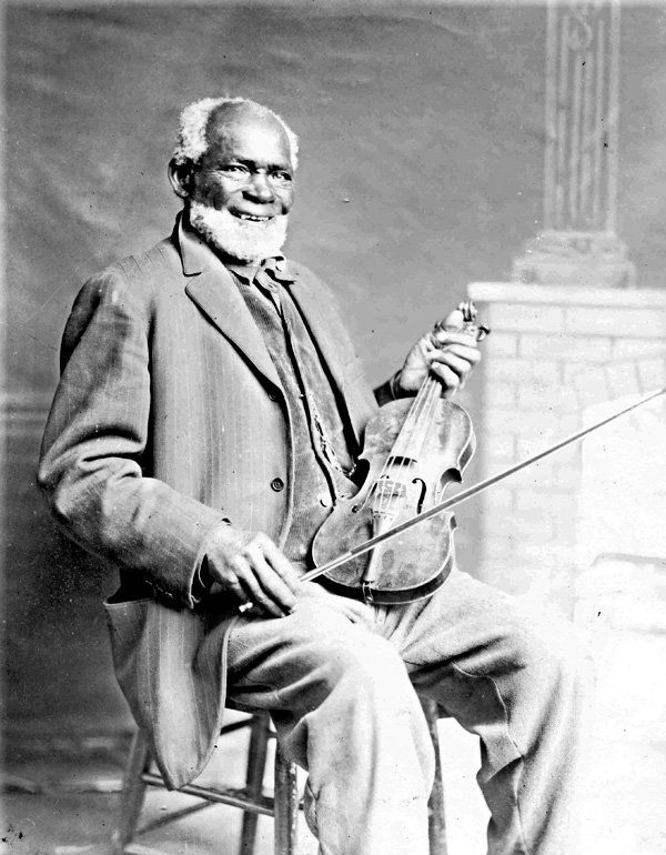 A smiling Louis Southworth sits in a wooden chair and holds his violin in one hand and bow in the other.