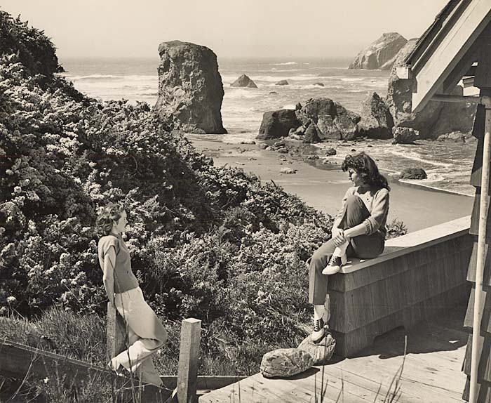 Two women relax on the porch of a cottage overlooking the ocean and impressively large rock formations.