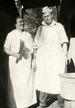 Woman and man standing. Woman holds a meat cleaver and both are dressed in aprons.