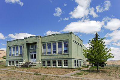 Two-story school building surrounded by dead grass. 1 small tree stands to the right. Blue sky with a few white clouds.