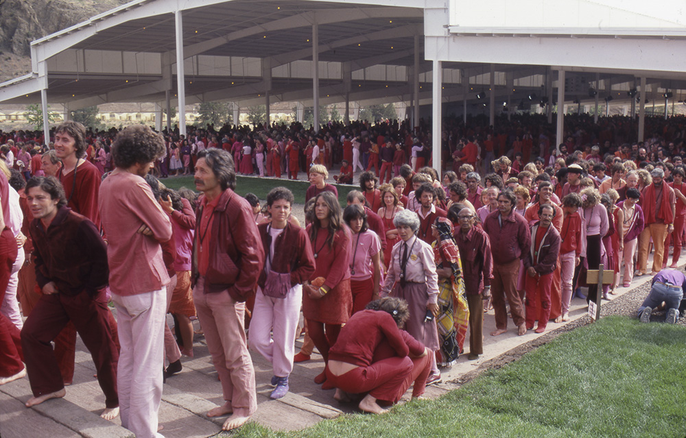 Hundreds of people, all dressed in redish or pink color clothing, stand in lines outside a building.