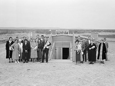 About 25 people, a few holding babies, dressed in suit or frock stand for a photo beside the opening to an underground shelter.