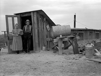Woman and man stand before a shed. Debris such as a card board box, metal barrels and wood planks lie about the area around.
