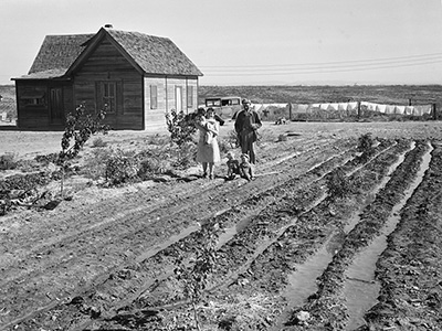 Woman holding baby, stands by a man and 2 children in a dirt field ready for planting. A small one-story house in the back. 