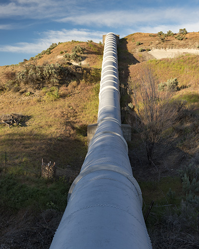 A metal pipe line extends up a hill and into the distance in an arid land with short grasses and bushes.