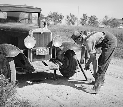 A man works a car jack set under the front bumper of a 1930s truck. A child stands behind him watching him work.