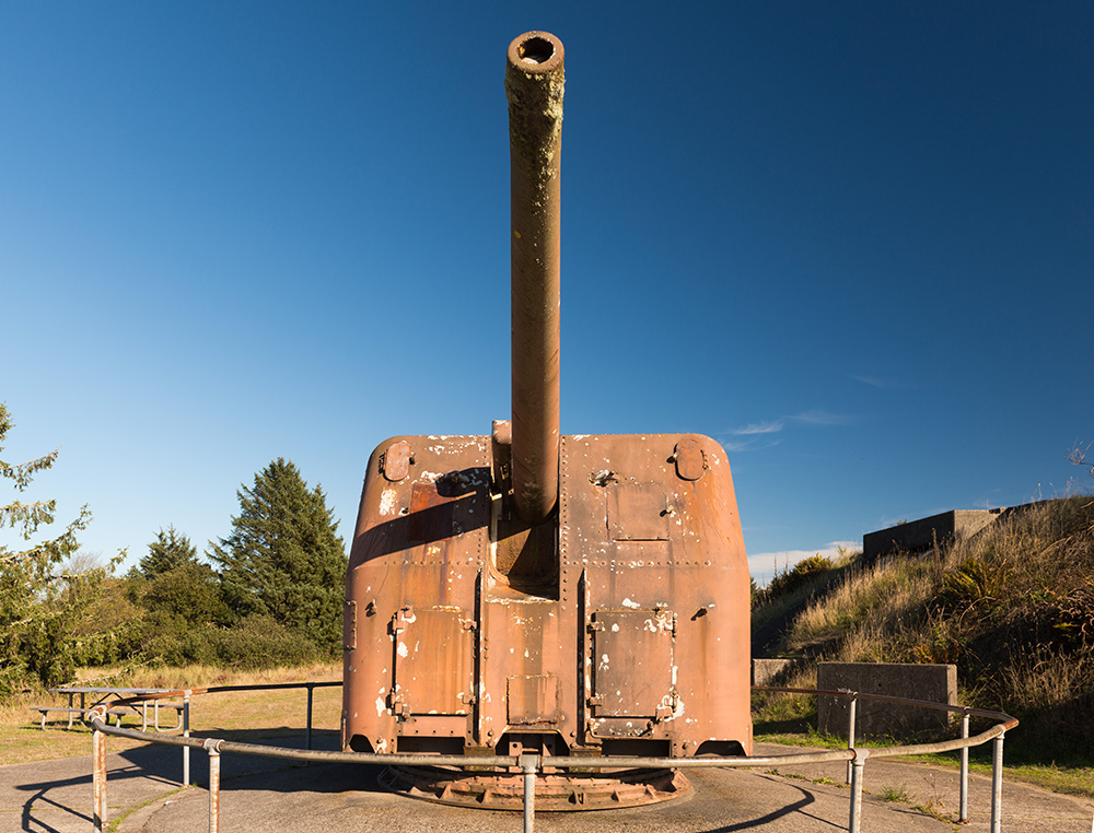 Metal artillery gun with a long barrel pointed toward the sky sits on a stone slap in a park. A picnic bench is nearby.