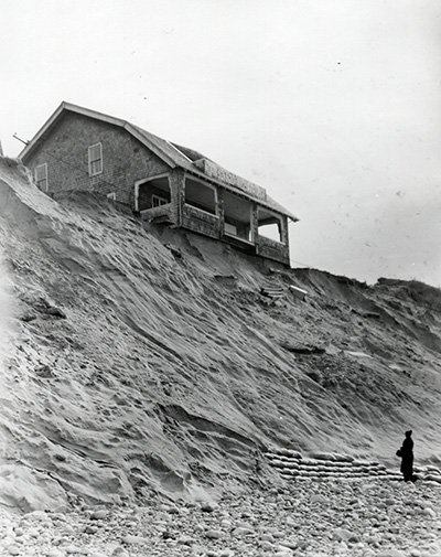 A two-story building with a porch rests at the edge of a cliff made of sand. It appears it will fall over the edge soon.