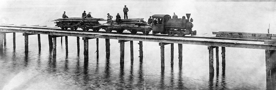 6 men stand on lumber slats loaded on rail cars. The rail line is built on wood pillars above the water. 