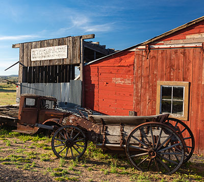 A rusted old truck and a wagon, falling apart with age sit in front of 2 dilapidated buildings in a field.