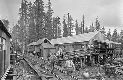 About a dozen men walk up a wooden ramp to get to a building used for meal times in the lumber company. A railroad track is near