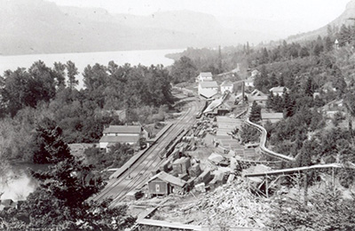 Photo of Bridal Veil taken from high atop a hill or mountain shows train tracks running along the river and buildings to east.