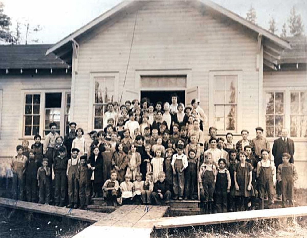 Approximately 70 people (students and teachers) stand in front of a single story building with tall windows. 