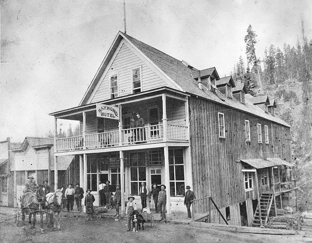 10 men stand on the porch of a 2-story building. a woman & girl stand on the balcony above them. 2 other men in a wagon.