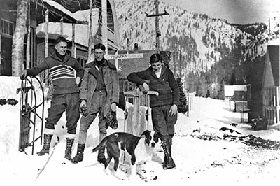 3 men & a dog stand in the snow. Two of the men have wood sleds. In the back a sign reads "Fountain ice cream." 