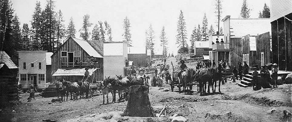 3 men with teams of 4 horses & wagon each stand in the middle of the road. About 15 to 20 people stand on porches of buildings.