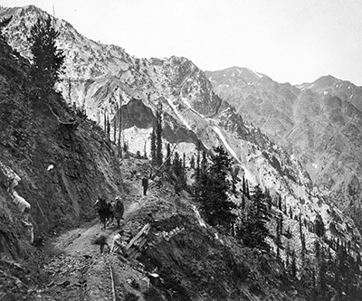 1 man sits on the edge of a dirt road while 2 others stand on the narrow road. 1 man standing has a horse. The area is rocky.