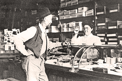Man in hat, shirt, vest & pants rests 1 arm on glass display case while woman behind counter weighs gold on scale. 