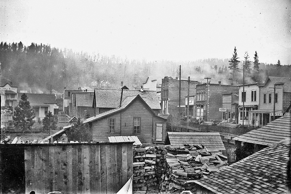 Black & white photo of Sumpter shows wood buildings close together & smoke rising from chimneys. Stacked firewood in foreground.