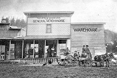 Men gathered around a general store in the late 1800s. 3 men sit on a wagon drawn by horses. 9 others stand on the porch. 