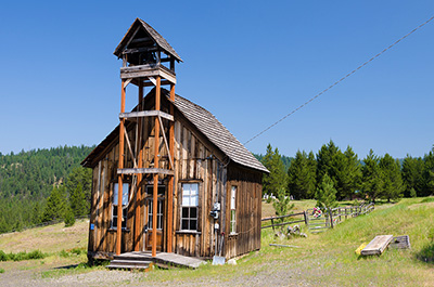 A wooden 1 room building with a bell tower over the front door. Blue sky and green evergreen trees with green grass surround it.
