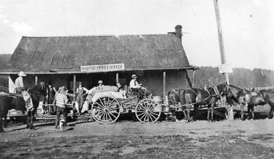 A wagon hitched to horses sits in front of a post office. Men stand around and some sit on horses or the wagon.