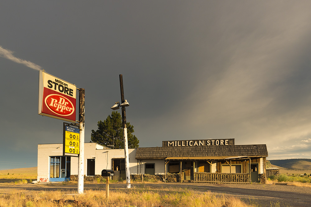 One-story road-side building stands under a stormy sky. The sign on the roof reads "Millican Store." A sign for Dr. Pepper in the foreground.