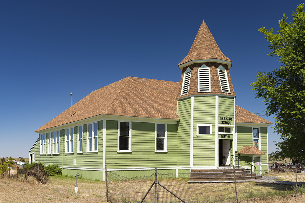 School is a one-story structure with a shake-covered hipped roof, boxed eaves, and shiplap siding. The entry is a two-story octagonal tower. 
