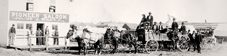 Some men sit in wagons drawn by horses while others stand on the wood boardwalk in front of a Pioneer Saloon. 