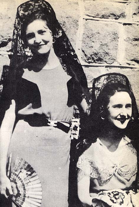 Two women, one sitting, one standing, in front of stone wall, smiling at camera. They wear dark veils on head. One holds a fan.