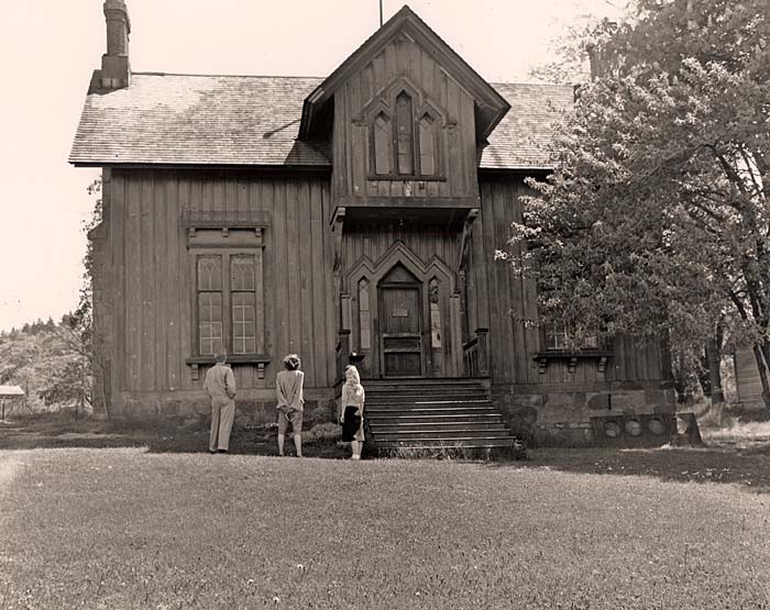 Three people stand in front of an old wooden building with a wide front door with a dozen steps leading up.