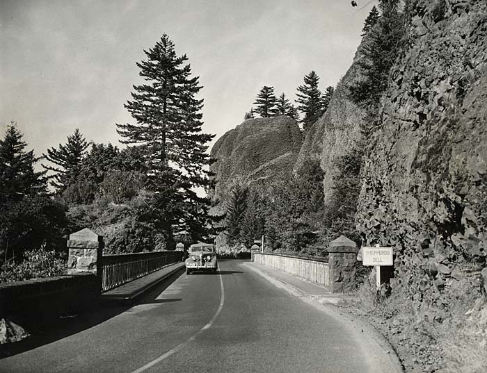 A paved 2-lane road leading over a stone bridge with a sign on the right that says "Sheppers Dell." A 1940s car drives toward us