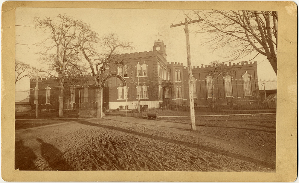 1871 photo of Oregon State Penitentiary shows a 2 story brick building with a clock tower in the center, surrounded by fence.