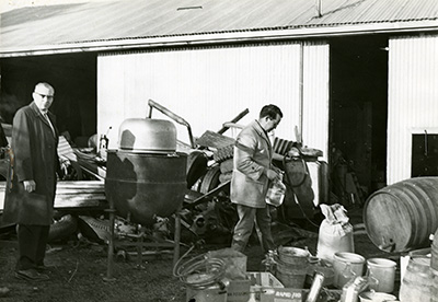 2 men are surrounded by still equipment such as barrels, metal pots, coils of copper, piping and other metal parts. 